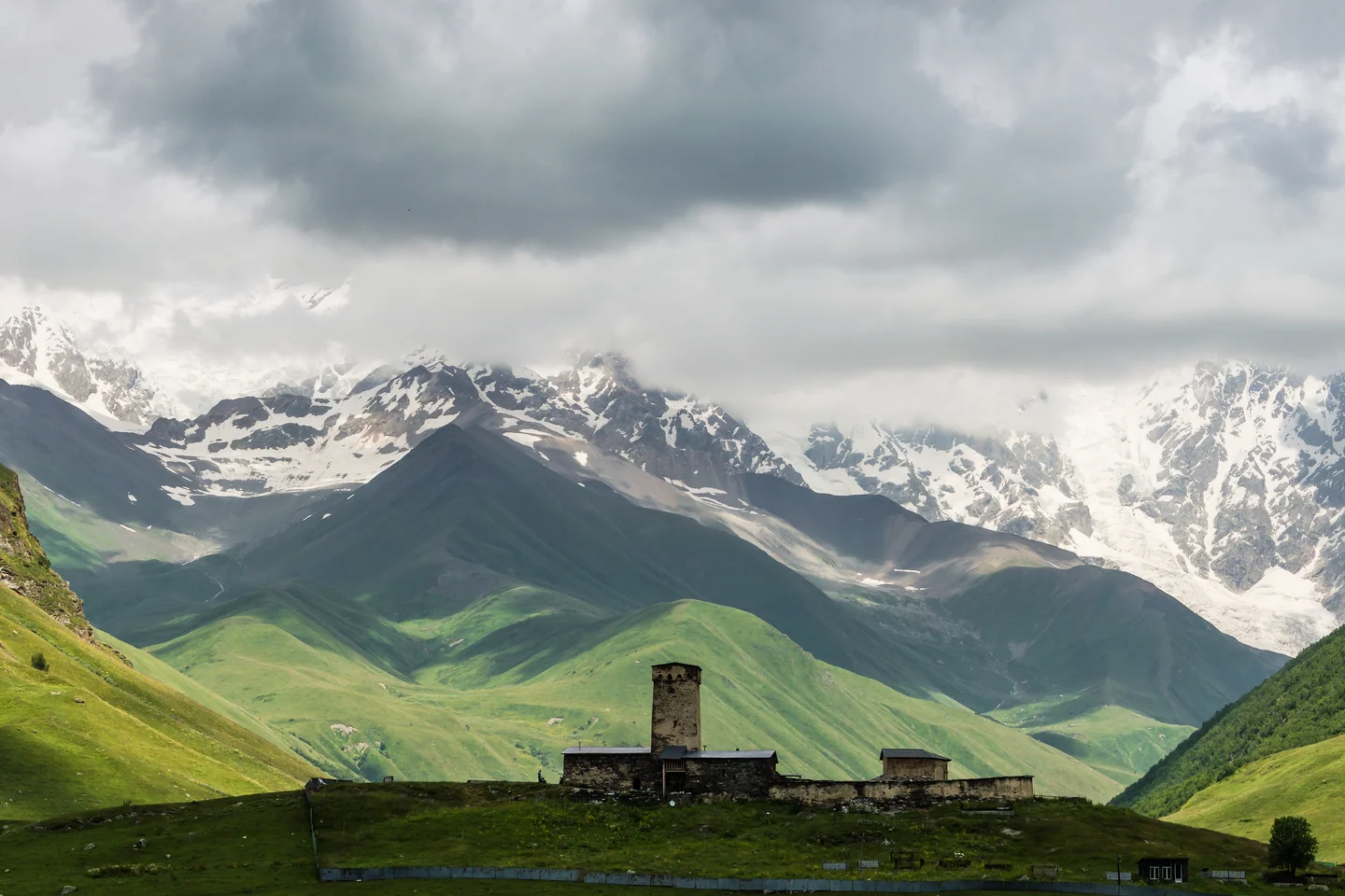 An ancient stone tower below sunlit Svaneti slopes and snow-covered Caucasus mountains
